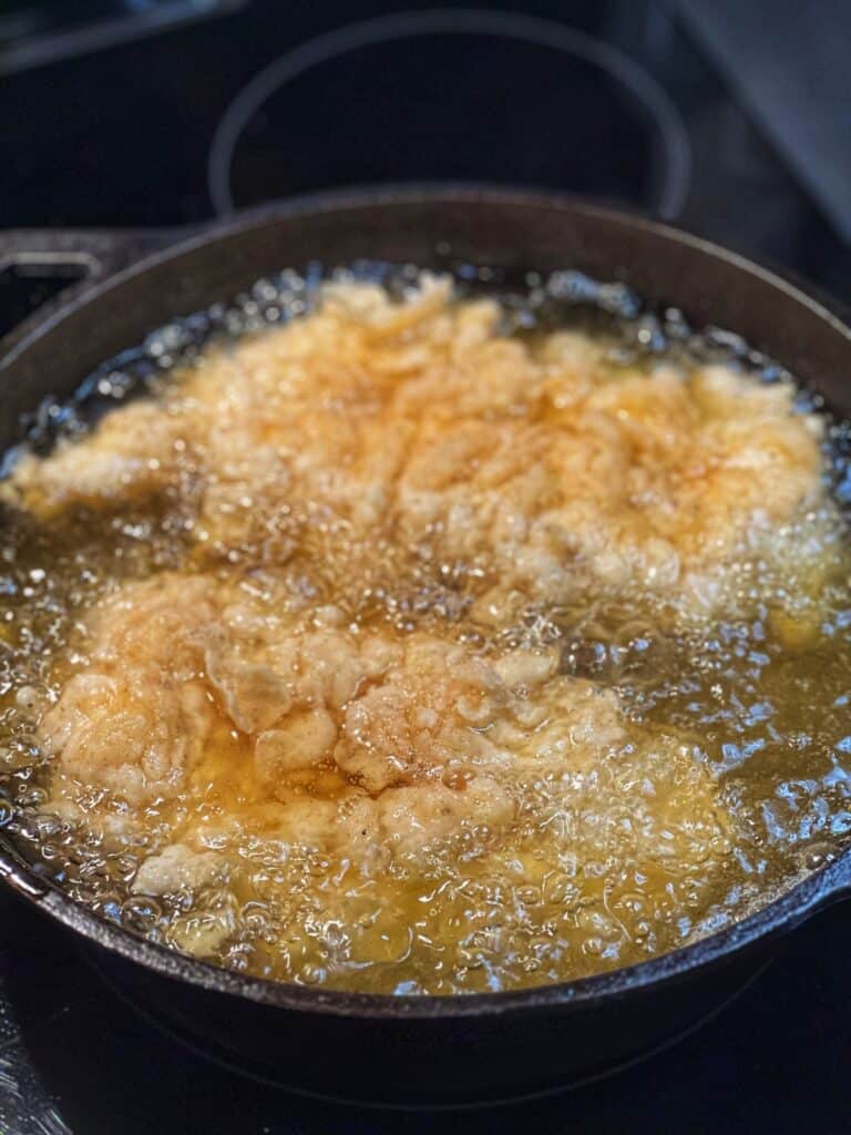 Two pieces of flour coated chicken starting to fry in hot oil in a cast iron pan.