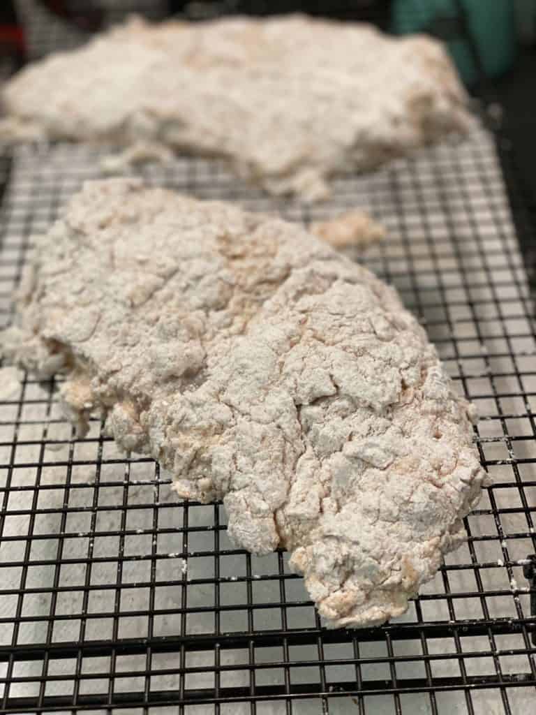 Two pieces of boneless chicken breasts coated in buttermilk and flour resting to dry on a cooling rack.