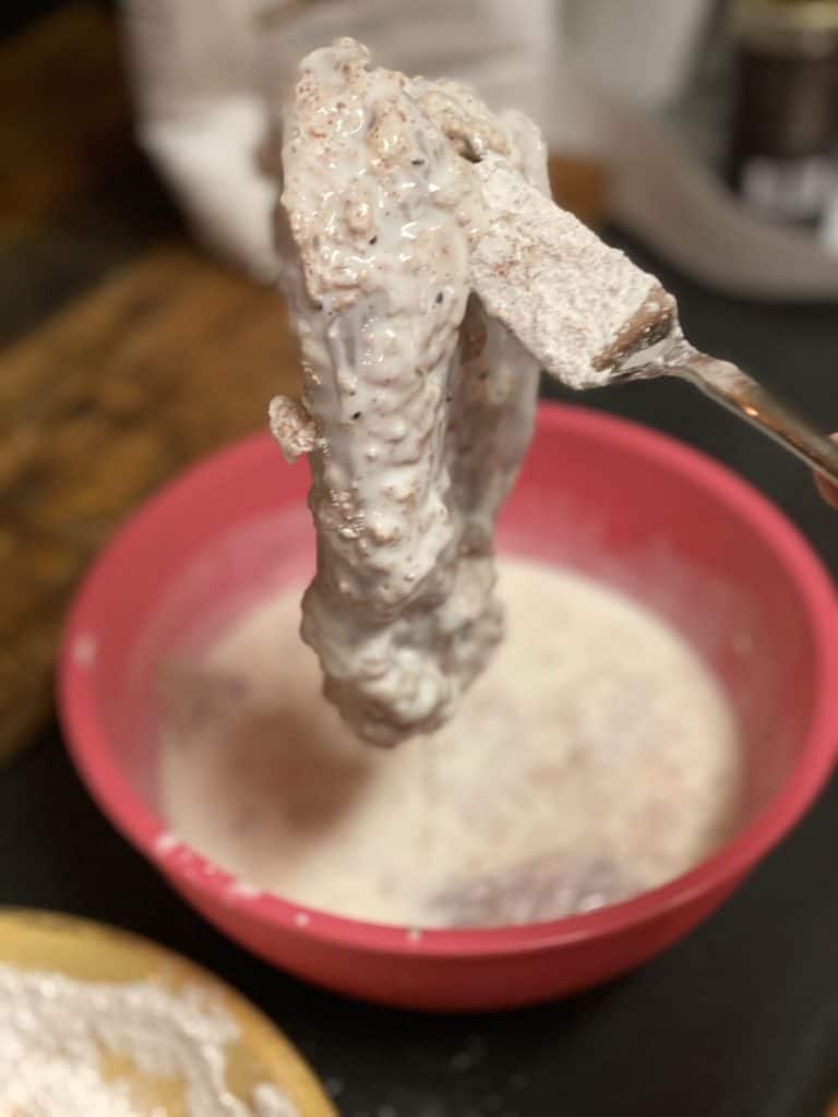 Holding a piece of chicken breast with a fork that is coated in buttermilk and flour.